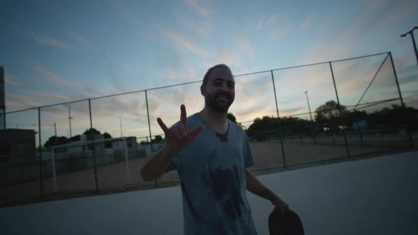 A man smiles and makes a hand sign towards the camera while standing outdoors in the evening
