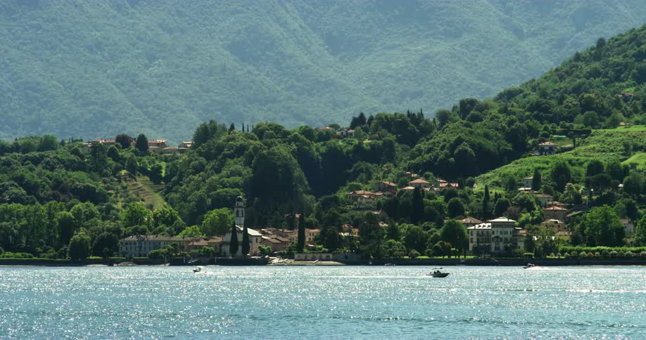 Motorboats move along sparkling water near shore of town with mist covered mountain in the background