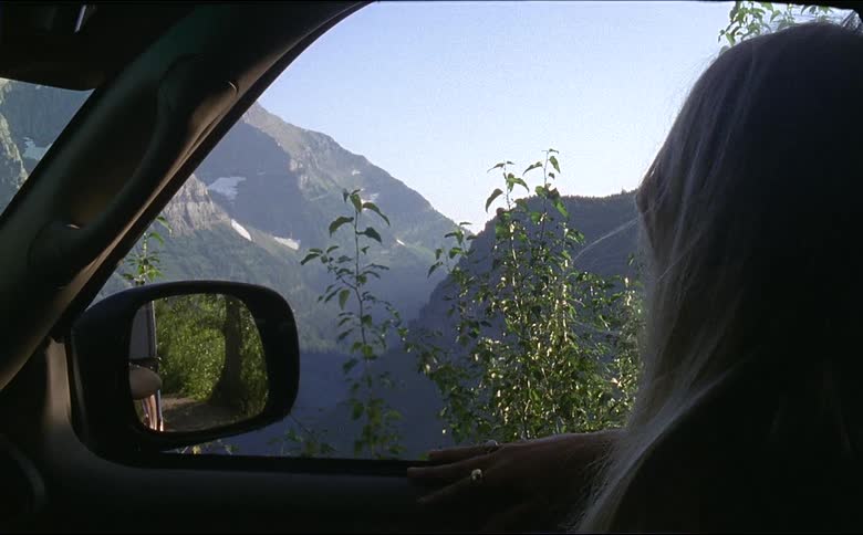 Young woman sitting on a car parked on the road looks at the mountains of glacier national park on 16mm film