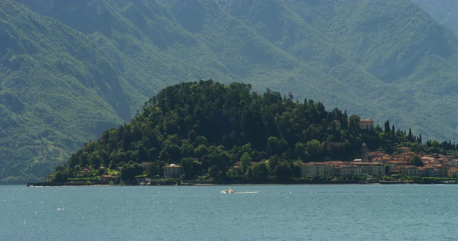 Small watercraft speeds by town on forest covered island with tall misty mountain towering in the background