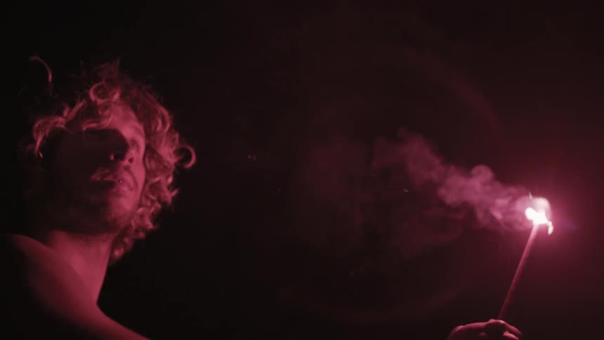 A shirtless man holds a flare at night