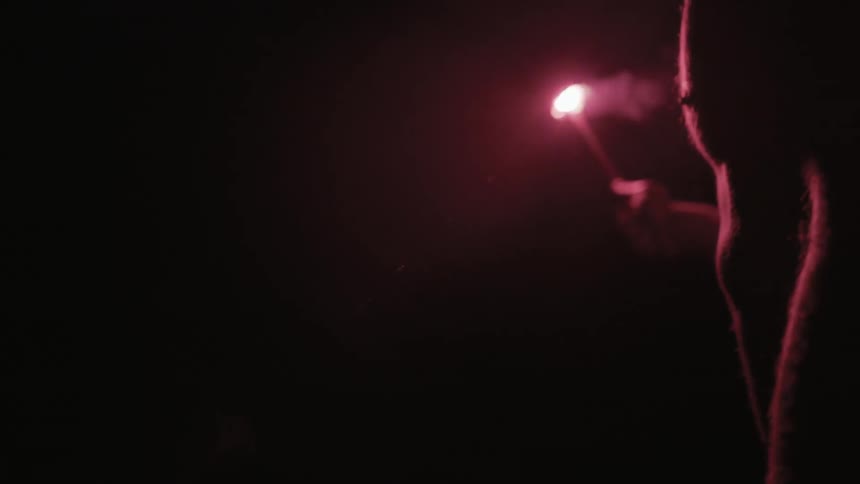 A bare chested man holds a flare at night