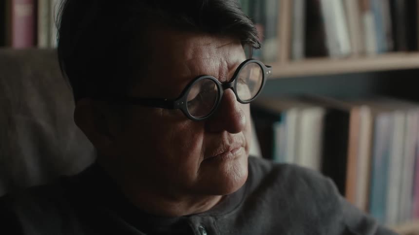 A man wearing glasses is sitting in a room with bookshelves on the wall and he is reading