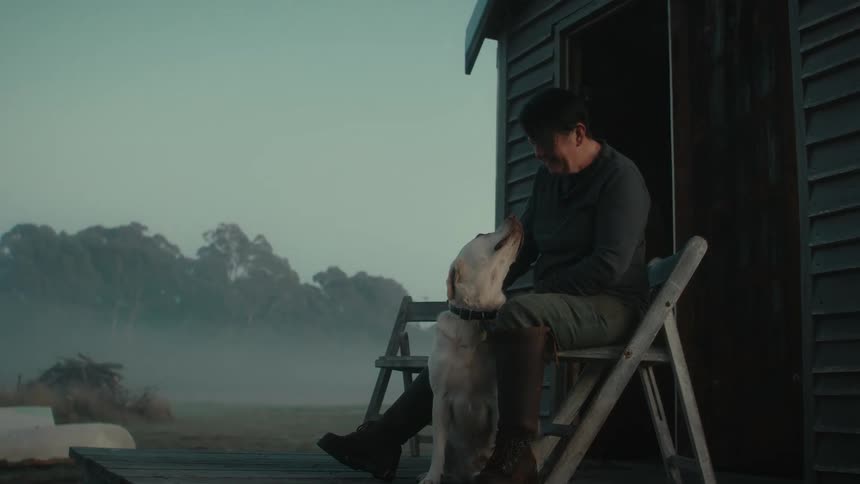 Woman pets her dog while sitting on a folidng chair in the porch of a small shack in the countryside