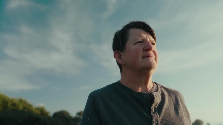Middle aged woman with short hair and gray sweatshirt looks into space smiling brightly in the countryside