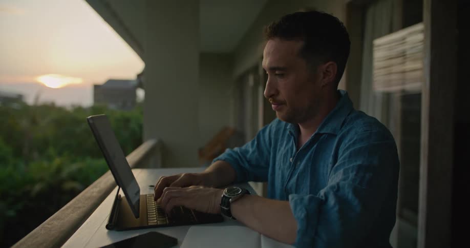Man works on a laptop over a tall desk on the balcony of an apartment on a tropical city