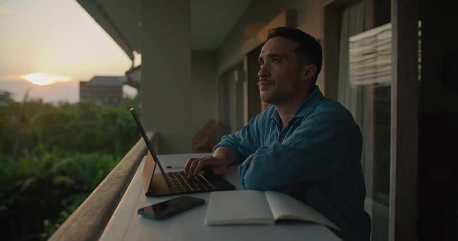 Man with blue shirt types on laptop keyboard in apartment balcony at sunset