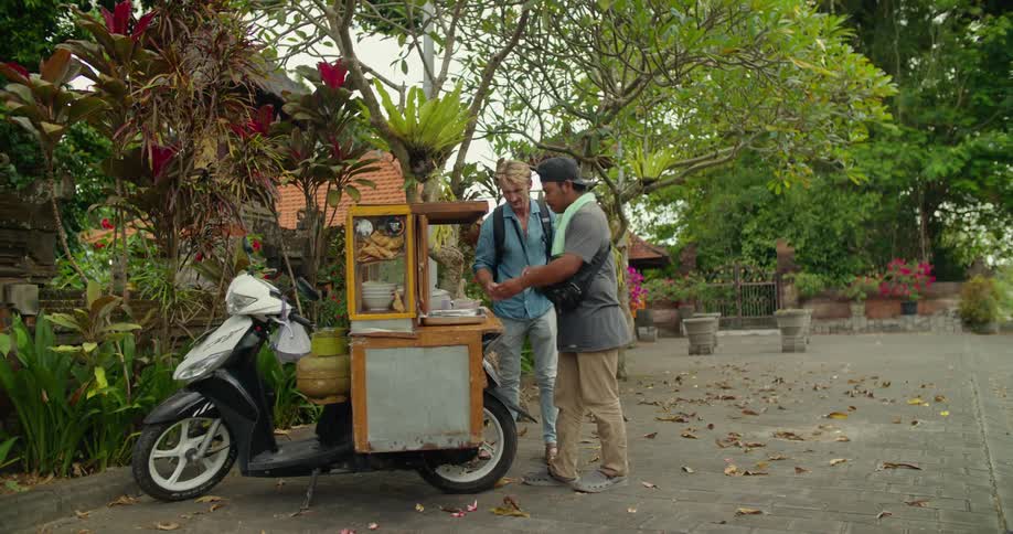 Blonde man with blue shirt jeans and backpack buys food from peddler in the street