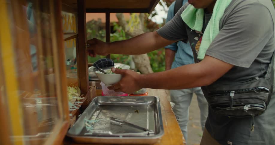 A man working at a food cart dishing up a bowl of food for a customer