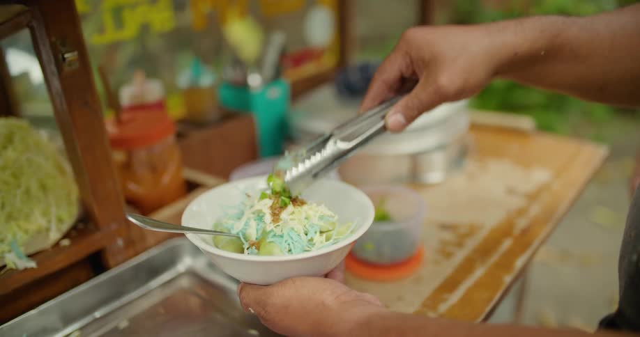 Hands using tongs add fresh green herbs as garnish to food in a bowl at a food cart