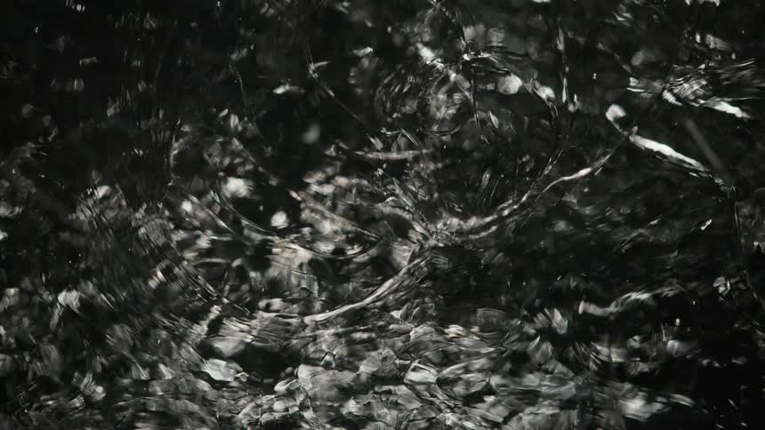 A clear substance reflects light as it ripples moving through a dark space like living glass