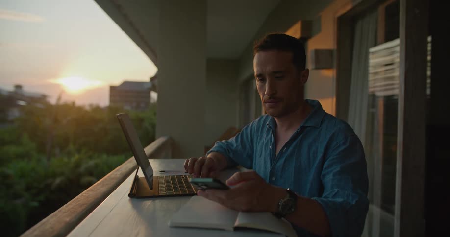A man is on his balcony looking at his cellphone and typing on his laptop