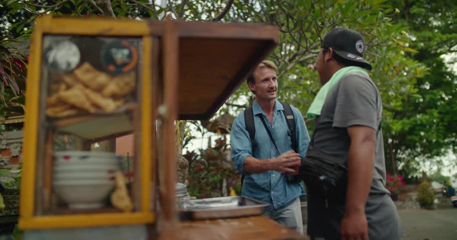 Tourist approaches a street food vendor and asks for a plate near a lush green garden