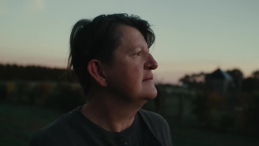 A person with a lined soft face and short hair standing outside deep in thought