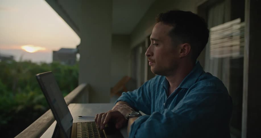 A man looks into the distance contemplatively before beginning to type on his laptop while sitting on the balcony of a building in the evening