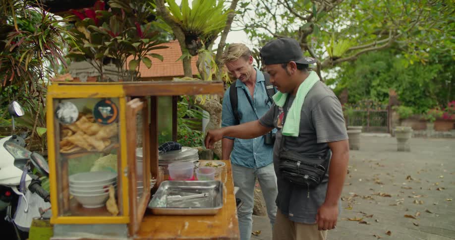 Blonde man with blue shirt white pants and backpack buys street food from peddler