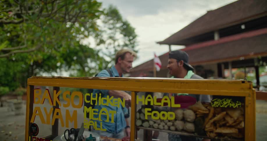 Blonde man with blue shirt and backpack buys food from peddler in the street