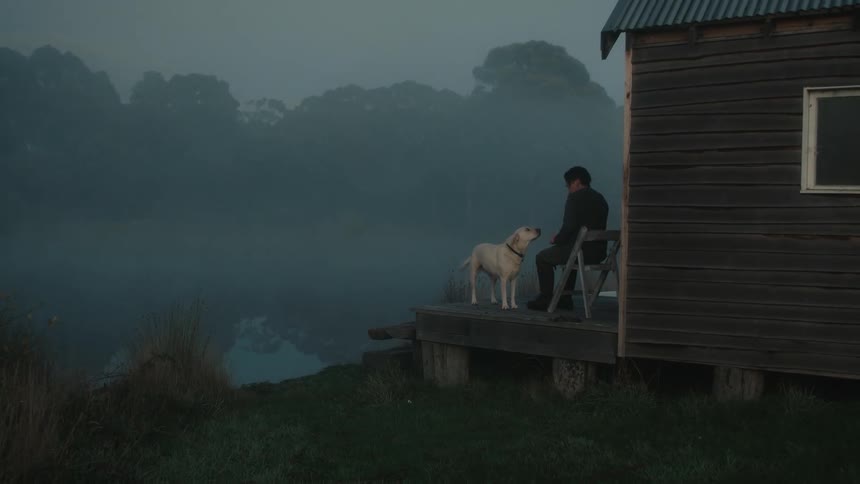 A person petting their dog while sitting on the porch of a lakeside cabin