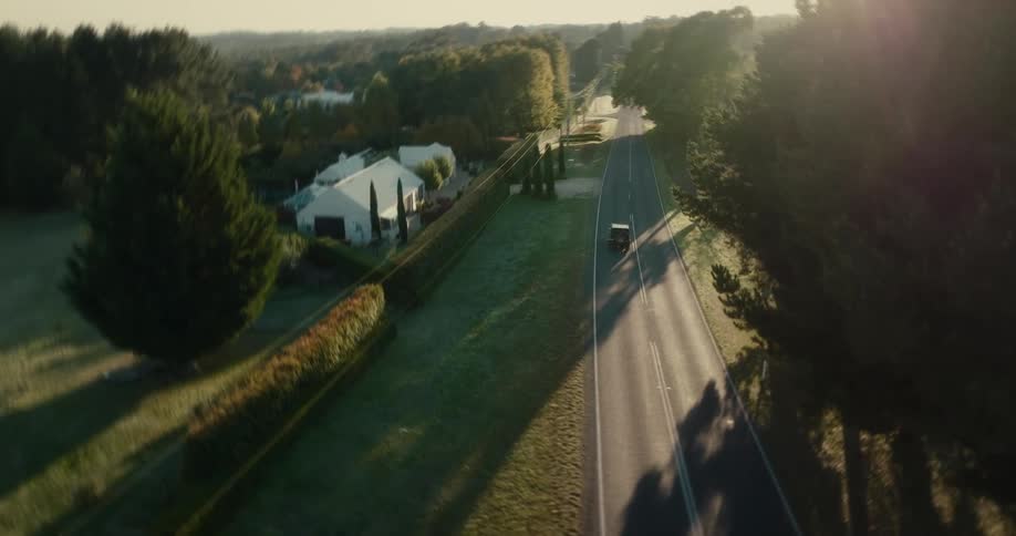 An automobile travels on a road lined with trees and houses as the sun shines brightly in the distance