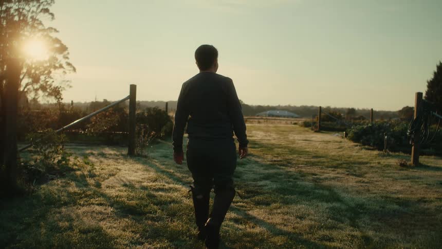 A woman wearing tall boots is walking on a grass path between plants and bushes