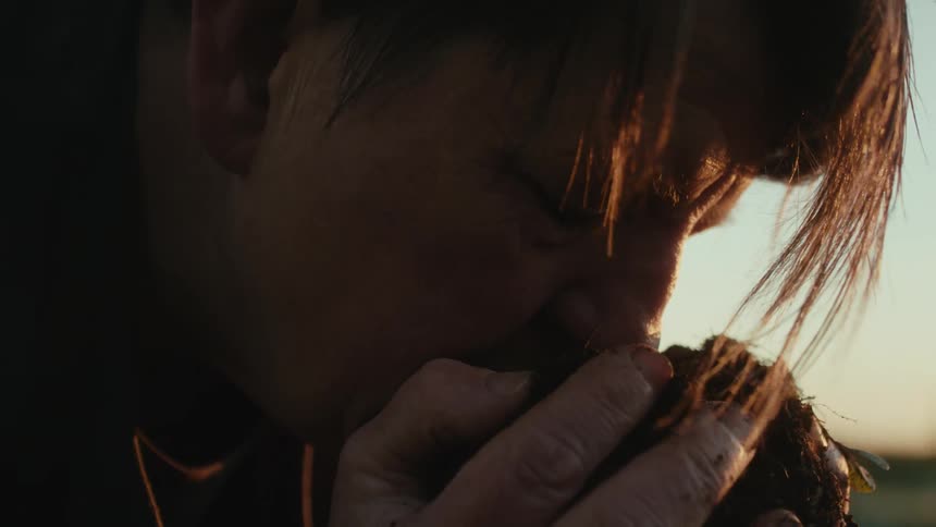 Middle aged woman with short hair sniffs soil in her hands in the countryside at sunset