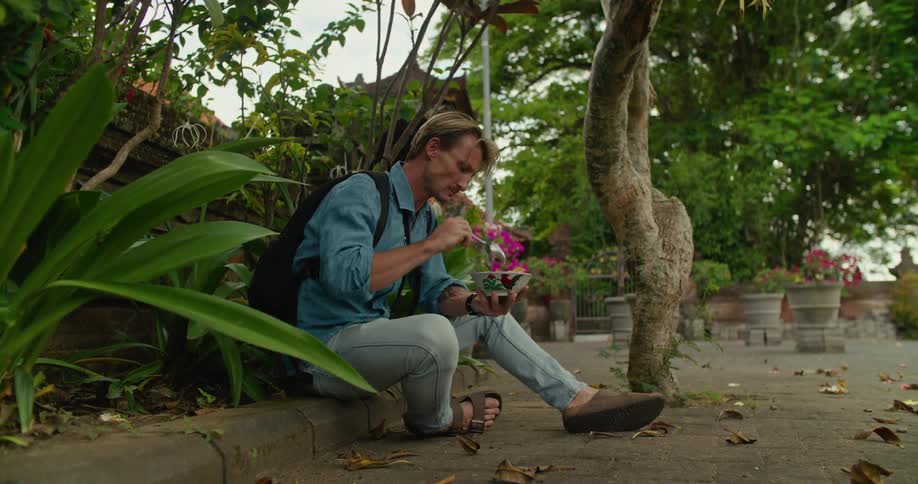 Blonde man with blue shirt jeans slippers and black backpack eats food from bowl in the street
