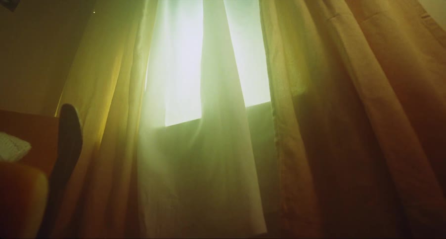 The wind blows a curtain through an open window and it moves in the breeze as sunlight shines in