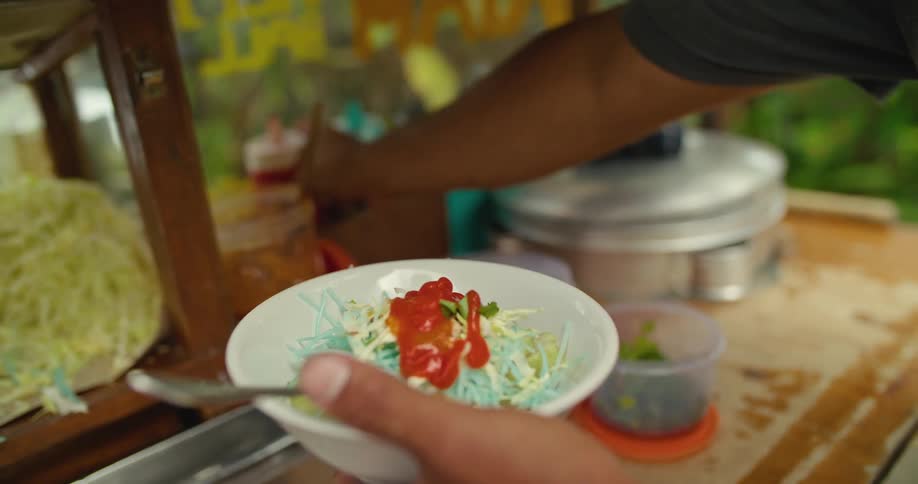 Man serving food on a street cart pours sauces on a dish