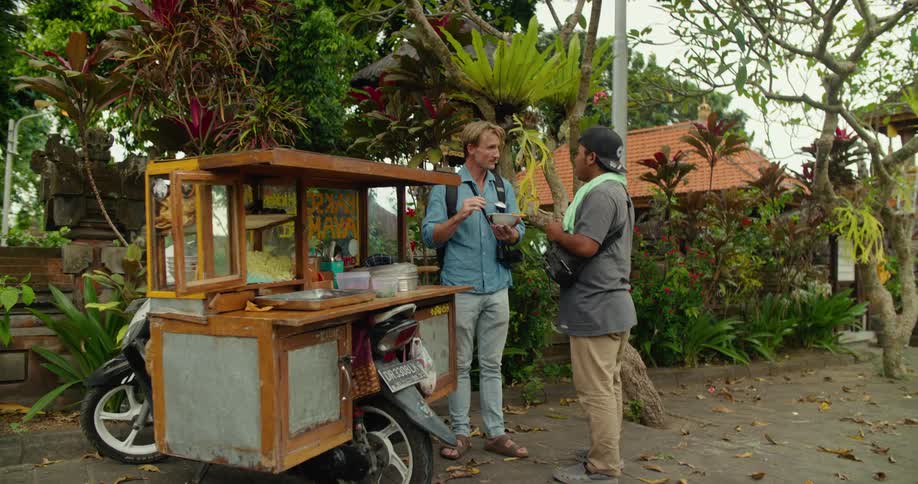 A man with a food cart on a motorcycle is talking to a man eating a bowl of food