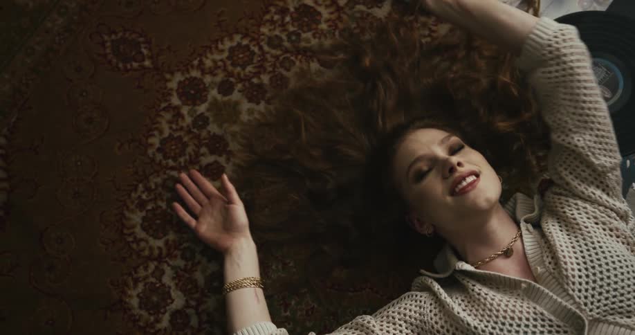 Woman with brown hair necklace and white button up jacket lies down on brown rug listening to music next to record discs