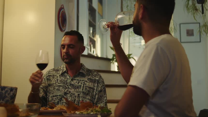 Two men are sitting at a table in a home drinking wine and eating food