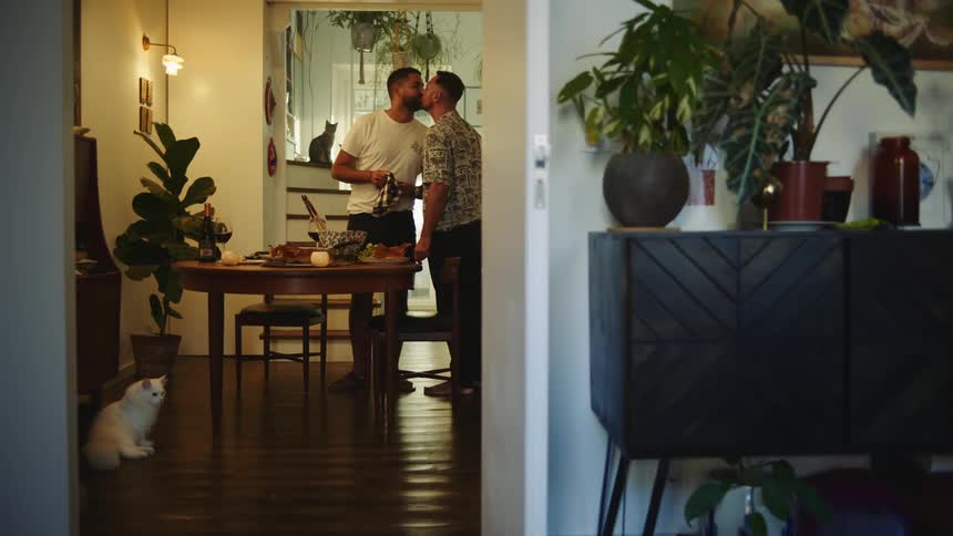 Two men kiss while picking up the plates from the dinner table at their cozy home
