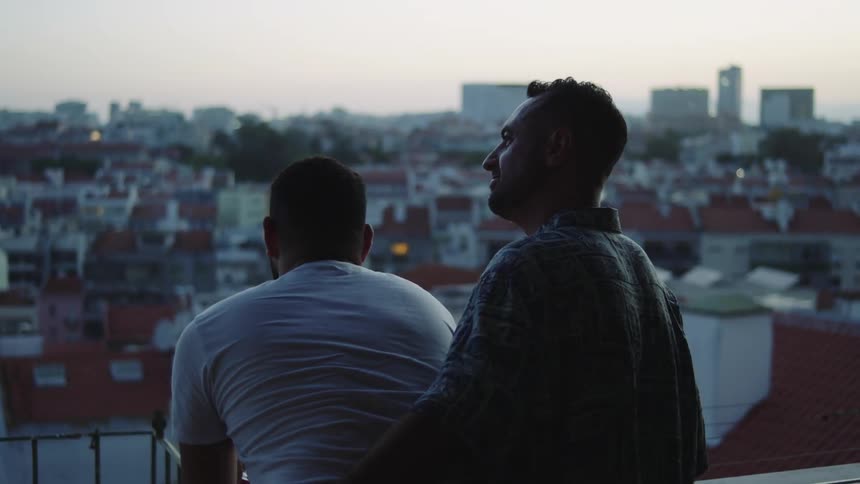Gay couple leaning on banister kisses and holds each other on balcony looking over neighborhood at dusk
