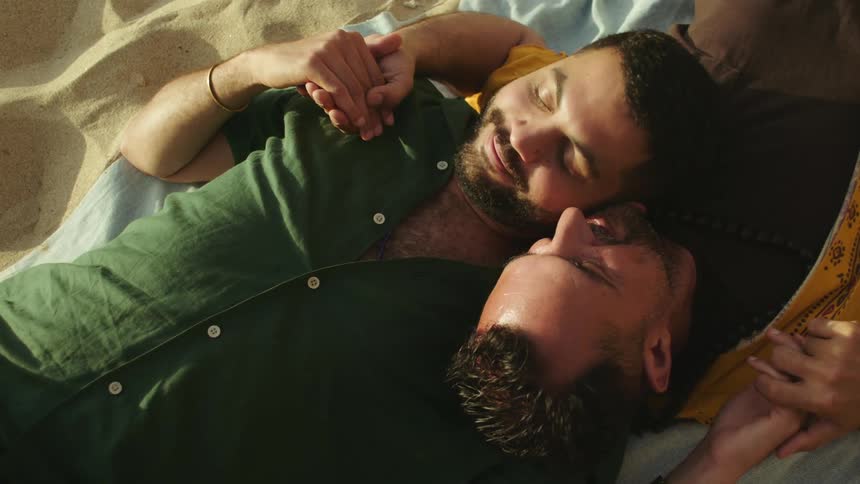 Two men are laying down and embracing on a towel on the sand