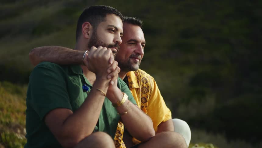 Two men are sitting outside together on a hill and one man has his arm around the other man and as they hold hands he kisses him on the cheek