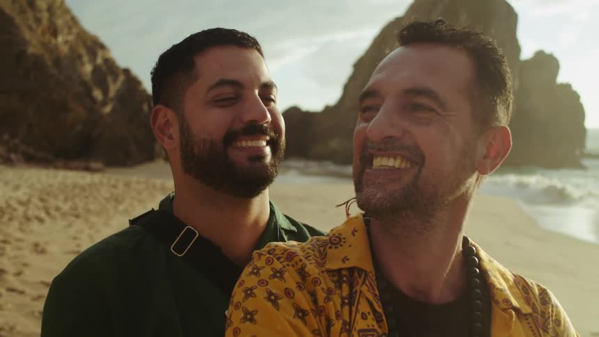 Two men are standing on the beach together hugging and smiling at each other then one man kisses the other on the cheek