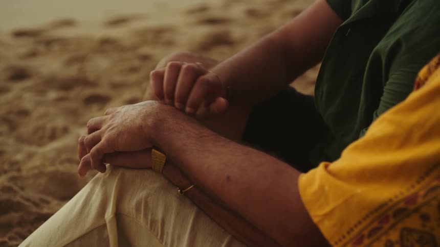 A man caresses his boyfriends arm and hand as they sit on the sand