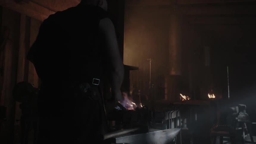 A large man tends to a tool near a fire inside a dim building