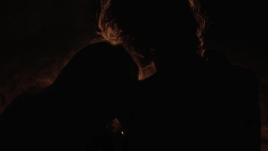 Two people lean against each other at night by a fire