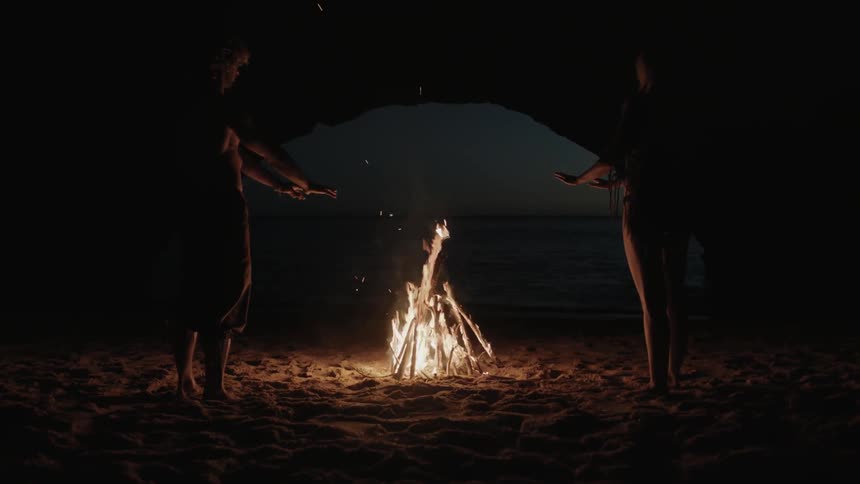 Two people stand by a fire in a cave near the ocean at night
