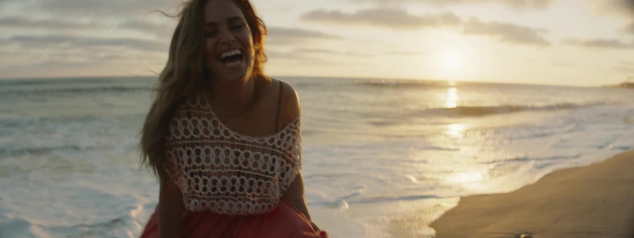 A woman runs away from the ocean waves smiling and laughing as her skirt billows around her
