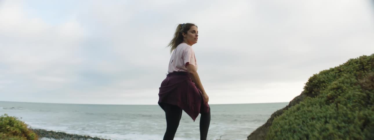 Woman with brown ponytail pink t shirt black leggings and crimson full zip hoodie around waist runs toward pebble beach on cloudy day