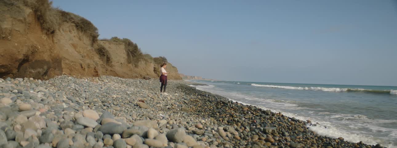 A woman walks on a stone covered shoreline and takes a picture as waves roll in