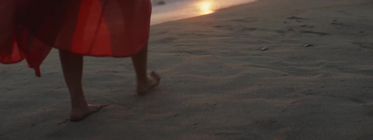 A woman walking barefoot in the sand with red skirt billowing around her legs at sunset