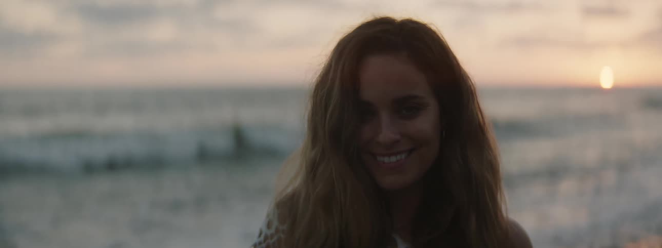 A woman is standing on a beach smiling as waves roll in to shore and the sun is on the horizon