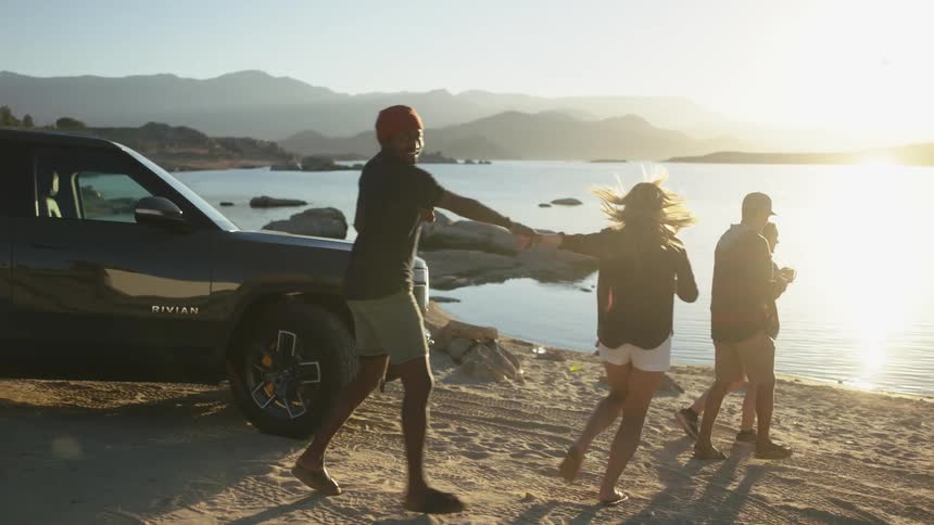 Two women with shorts and shirts pull their boyfriends hands from standing next to black pickup truck to the beach at sunrise
