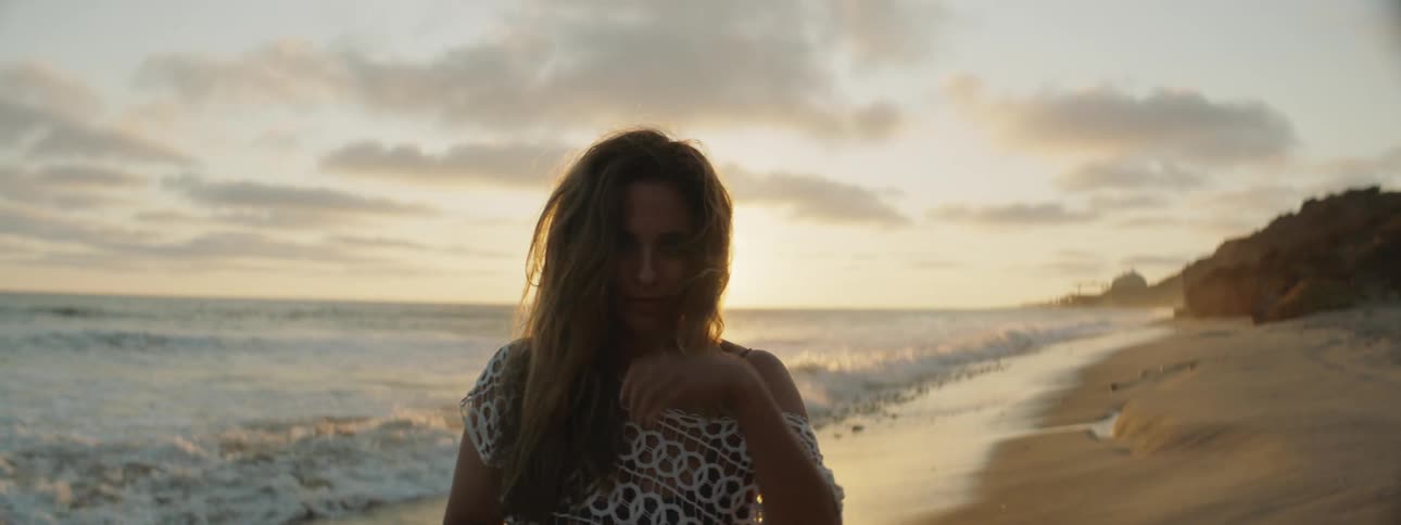 A young woman on the beach brushes her hair off her face as waves roll in