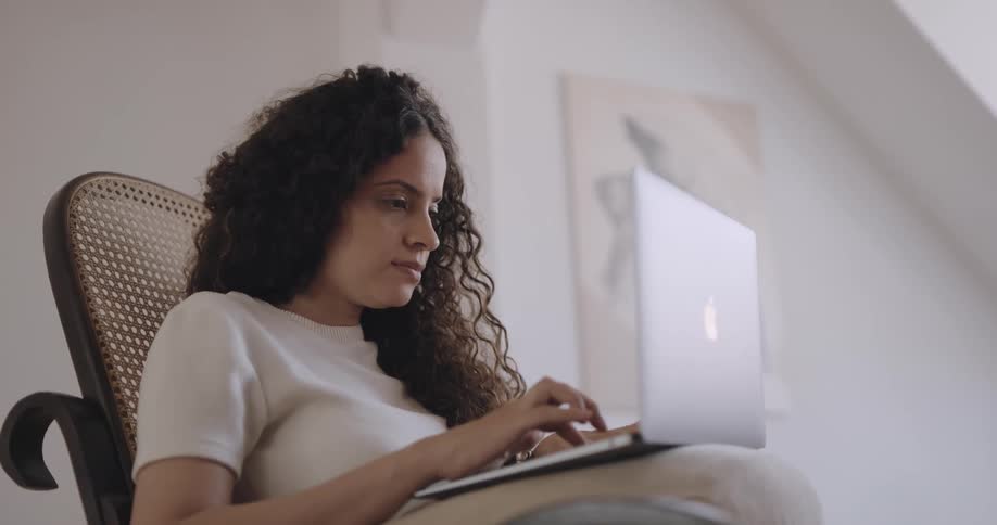 Woman with curly hair and white shirt sits in armchair using laptop in the living room