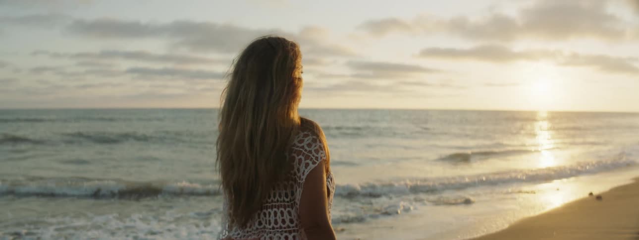 Young woman wearing a long dress walks on the sand next to the coast at sunset