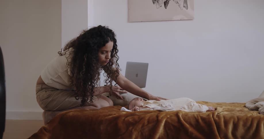 A woman is sitting on the edge of a bed with a laptop computer as she tries to comfort her baby lying in the bed